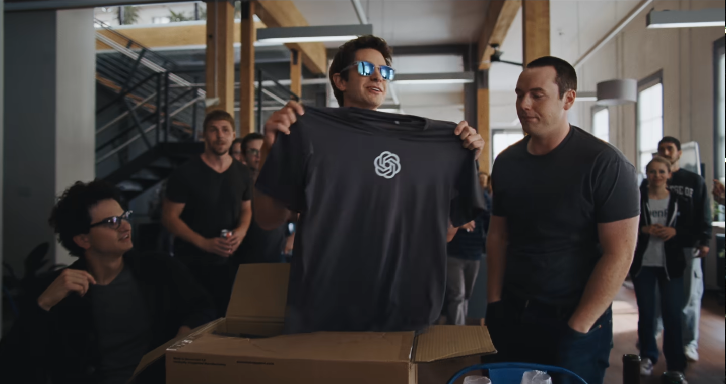Artificial Review: A Scathing Look at OpenAI [NYFF 2026] A man surrounded by co-workers holds up a t-shirt